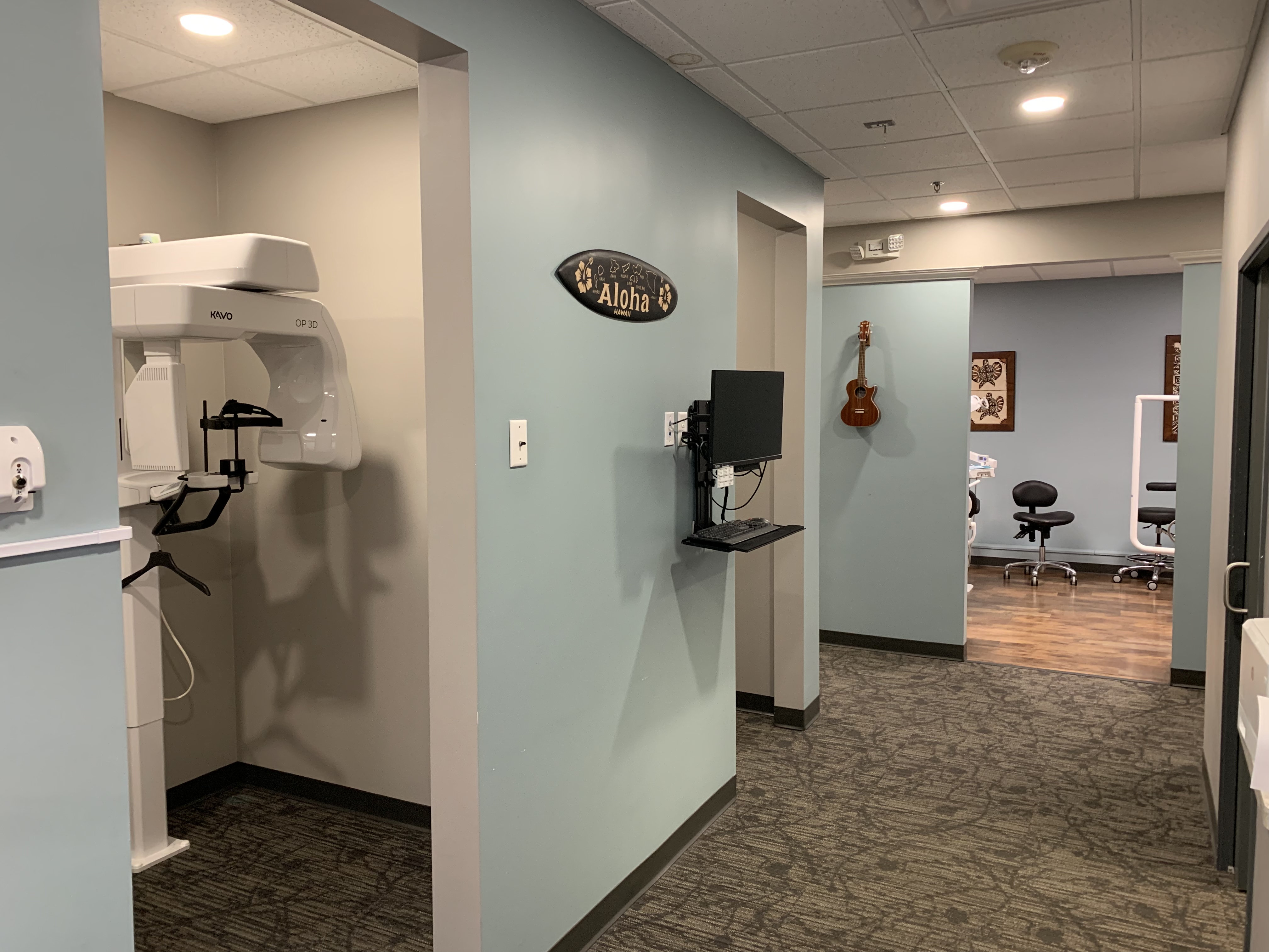 Interior of a dental office with a panoramic dental x-ray machine on the left, a computer station, and a ukulele hanging on the far wall with a sign that reads "aloha" above it | Pleasant Dental