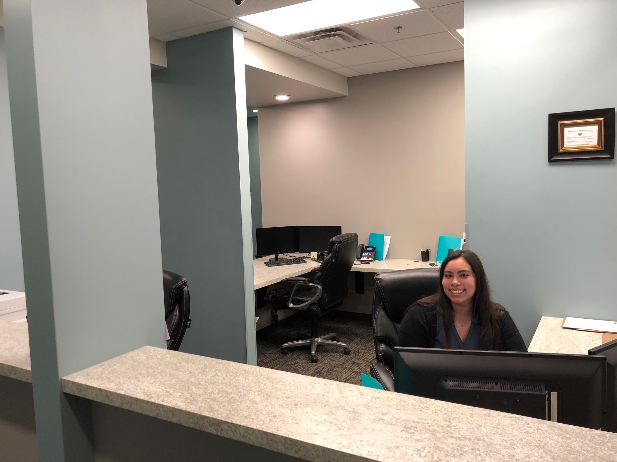 A woman smiling at the reception desk in a modern office with computer workstations and blue chairs in the background | Pleasant Dental