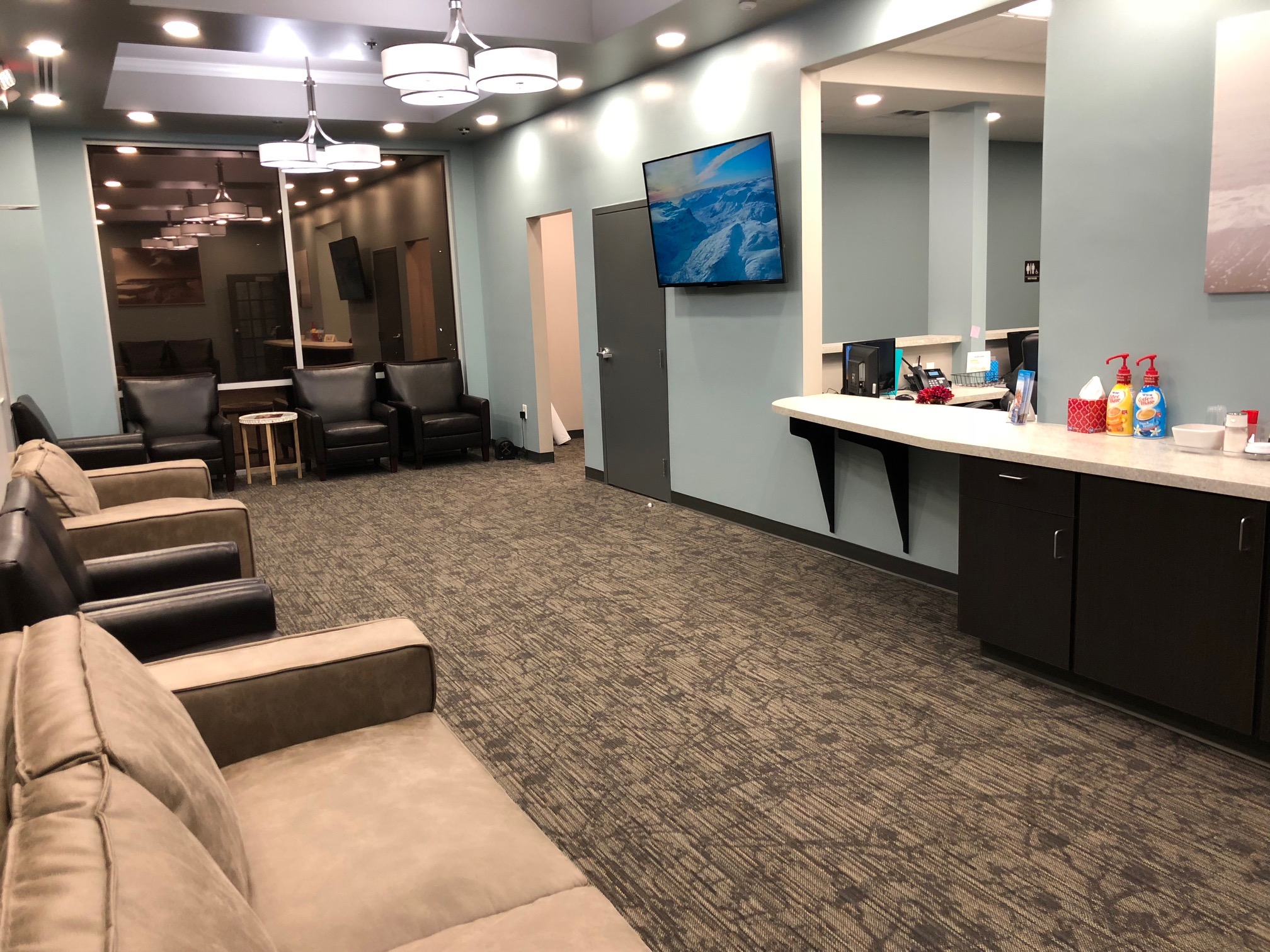 A modern office break room with dark leather chairs, a blue accent wall, mounted tv, kitchenette with snacks on the counter, and patterned carpet flooring | Pleasant Dental