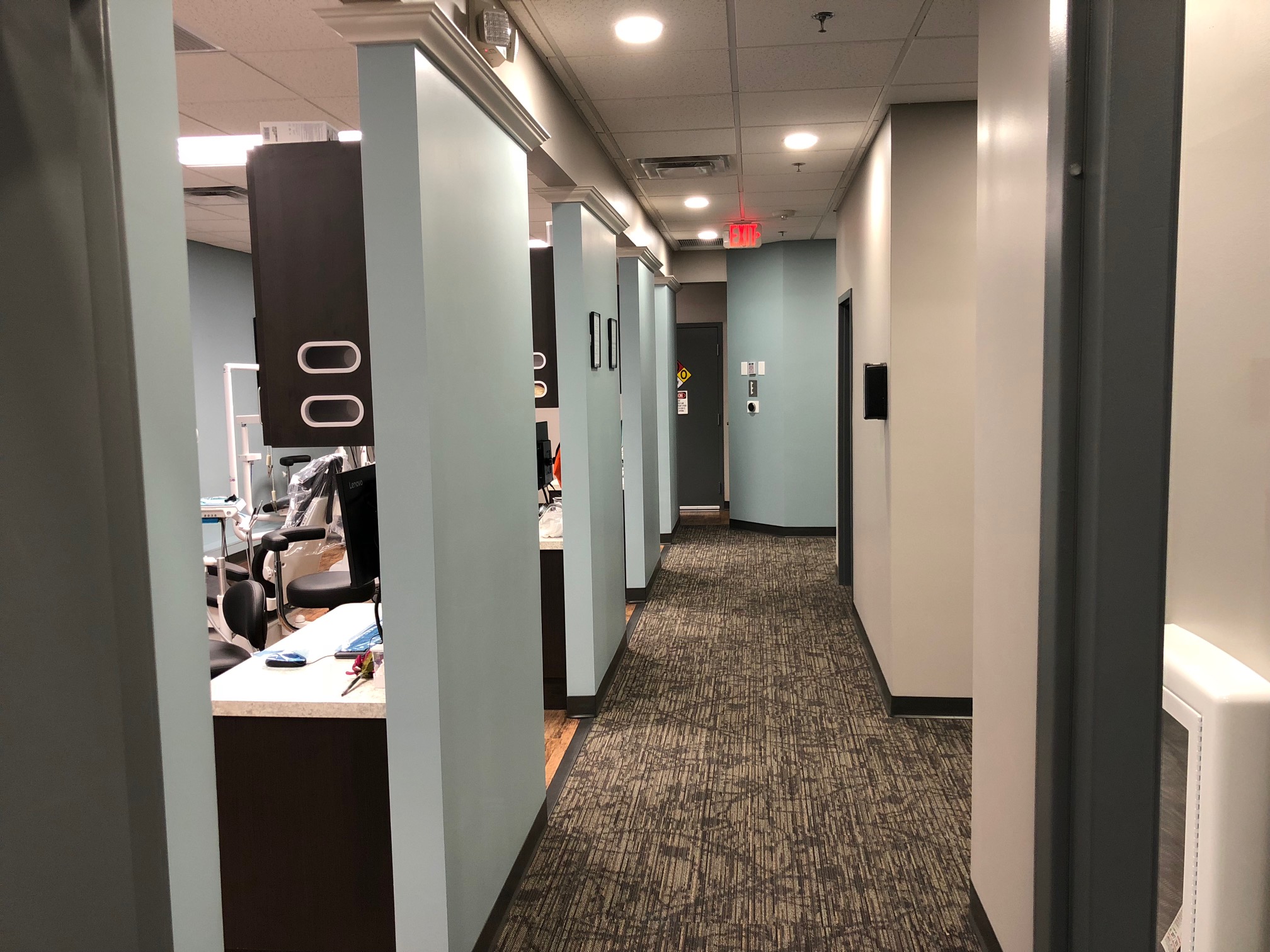 A hallway in a modern office or clinic with partial views into rooms through open doors. the corridor features teal walls, decorative mirrors, and a dark carpeted floor | Pleasant Dental