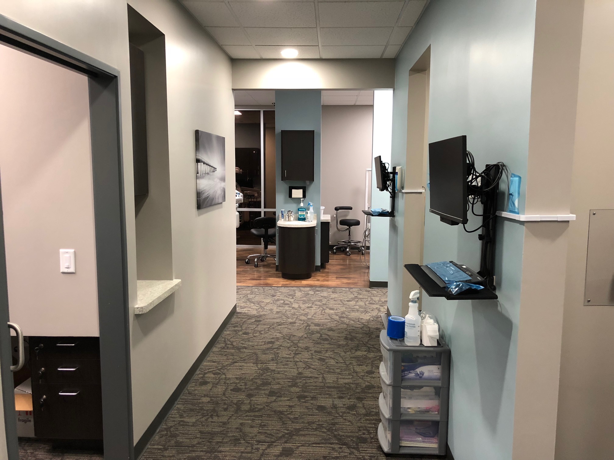 A modern dental clinic corridor featuring an open door leading to a treatment room with a dental chair, monitors mounted on walls, and framed artwork. the floor is carpeted, and the walls are light gray | Pleasant Dental