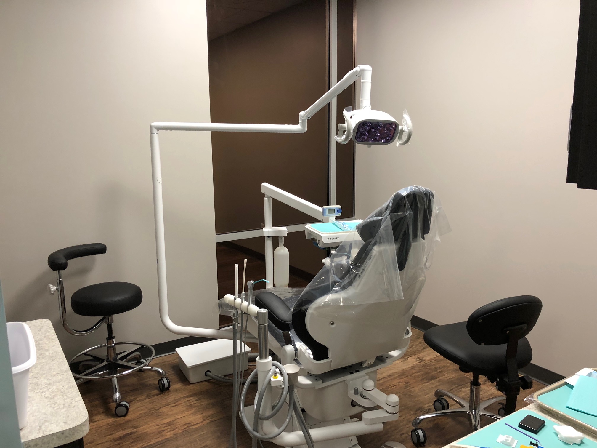 A modern dental clinic room featuring a dental chair covered with a protective plastic sheet, surrounded by various dental equipment, tools, and two stools | Pleasant Dental