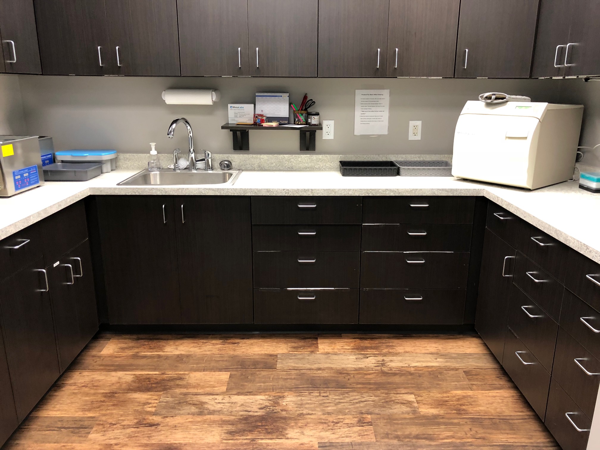 A modern, clean kitchen corner with dark wood cabinets, a stainless steel sink, and various appliances on a speckled countertop | Pleasant Dental