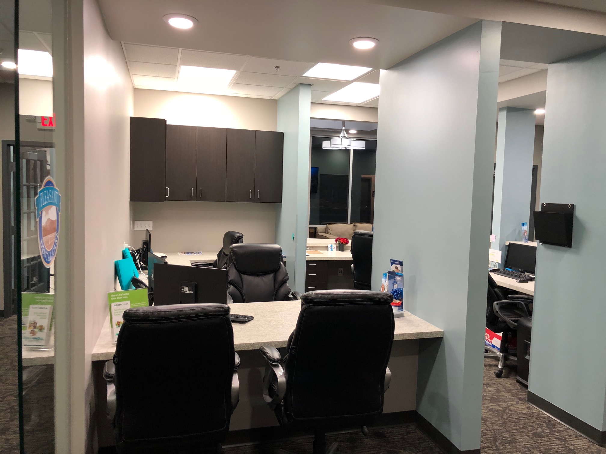 A modern office space featuring a reception area with two black office chairs, a desk with a computer, and cabinets above. there's a window offering a nighttime view, and a hallway leading further inside | Pleasant Dental